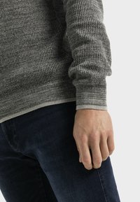 Harmaa ribbineulottu neule, jossa on vaaleampi, kontrastinvärinen ribbineuloshelma, päällä tummansiniset farkut. Neuleessa näkyy tekstuuri ja istuva muotoilu.