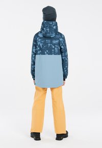 Giacca impermeabile blu scuro con parte superiore a fantasia e parte inferiore blu chiaro. Abbinata a pantaloni gialli e stivali neri, completata con un berretto grigio.
