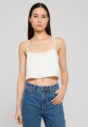 Ung kvinde iført en hvid ærmeløs cropped top med knyttede stropper og højttaljede blå jeans, stående mod en ensfarvet baggrund.