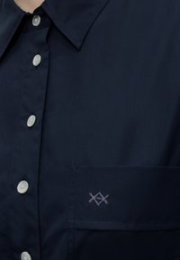 Κοντινό πλάνο μπλε πουκάμισου με άσπρα κουμπιά και μικρό κεντημένο λογότυπο πάνω από την αριστερή τσέπη στο στήθος.