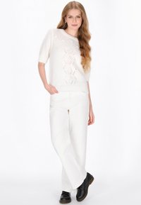 Biały sweter dzianinowy z krótkim rękawem, z otwartym wzorem; połączony z białymi spodniami w szerokim kroju i czarnymi butami sznurowanymi.