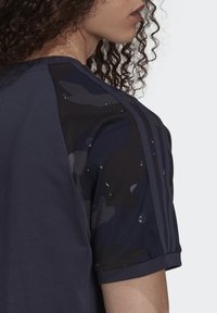 Marineblaues Sport-T-Shirt mit strukturierten Camouflage-Ärmeln, das drei vertikale Streifen an der Schulter aufweist. Aus Baumwollmischung.