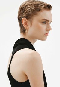 Top negro sin mangas con un diseño drapeado, combinado con pendientes de aro dorados ornamentados que presentan patrones geométricos, lucidos por una modelo de perfil.