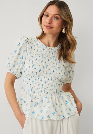 Kvinde med bølget hår, iført en hvid smock-bluse med blåt blomsterprint, pufærmer og guldringearringe, hænderne i lommerne.