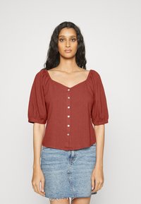 Blusa de cor ferrugem com mangas curtas bufantes, decote amplo e detalhes de botões na frente. Usada com uma saia de jeans clara.