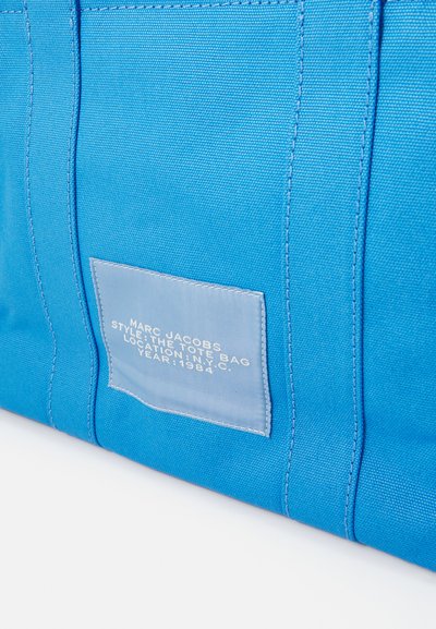 Sac fourre-tout en toile bleu avec des accents cousus, présentant une étiquette bleu clair avec un texte précisant la marque, le style, le lieu et l'année.