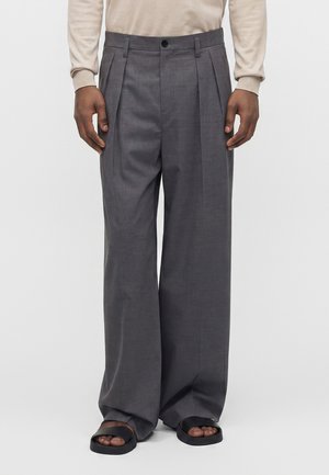 Homme portant un pantalon gris à jambes larges avec des plis devant, une chemise beige à manches longues et des sandales noires à bouts ouverts, debout devant un fond uni.