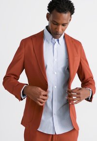 Blazer texturé couleur rouille avec un revers en pointe, associé à une chemise à rayures bleu clair. Les détails incluent une poche poitrine unique et des poignets à boutons.