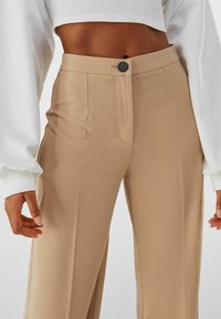 Beige högmidjade byxor med en slät textur, en knappstängning och sidofickor, kombinerat med en vit långärmad topp.