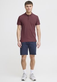 Bordeaux polo skjorte med krave, tre knapper og logo detalje, parret med marineblå shorts samt hvide sportssokker og sko.