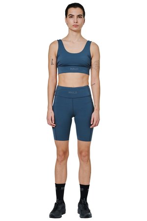 Kvinde model med mørkt hår iført blå sports-bh og cykelshorts, sorte sokker og sorte sportssko, stående vendt fremad på hvid baggrund.