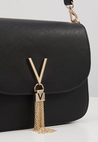 Bolso negro con una superficie texturizada, que presenta un logotipo en oro en forma de "V", un cierre metálico y un detalle de borla en cadena dorada.