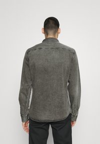 Grå denimskjorta med spetskrage och knäppta manschetter, med en strukturerad yta och rundad nederkant, visas bakifrån.