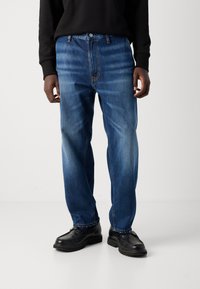 Man in blauwe vervaagde rechte spijkerbroek, zwart overhemd met lange mouwen en zwarte leren schoenen, staand op een witte vloer.