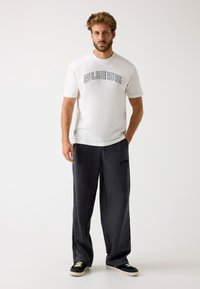 T-shirt bianca a maniche corte con logo "GUESS", abbinata a pantaloni della tuta grigio scuro con strisce laterali e logo, completata da sneakers nere.