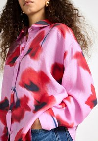 Růžová košile na knoflíky s červenými a černými květinovými vzory, lehká tkanina, volný střih a límec. Nošeno s modrými džínami.