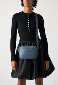 Borsa a tracolla in pelle blu con una texture liscia e dettaglio del logo. Abbinata a un abito nero a coste e gonna a campana.