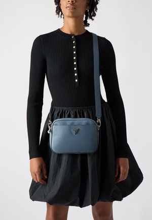 Borsa a tracolla in pelle blu con una texture liscia e dettaglio del logo. Abbinata a un abito nero a coste e gonna a campana.
