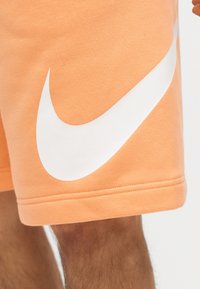 Broskyňové šortky s veľkým bielym logom Nike Swoosh na stehne, nosené osobou s viditeľnými chlpmi na nohách.