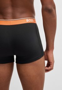 Boxers noirs avec un waistband orange portant le nom de la marque "BOSS" en blanc. Le tissu semble lisse et extensible.