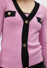 Rosa ribbad cardigan med svart kant, med guldknappar och två bröstfickor. Texturen är jämn, med detaljer i vertikala ränder.