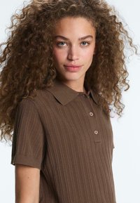 Femme aux longs cheveux bouclés portant un polo marron à manches courtes côtelé, face légèrement de profil avec une expression neutre.