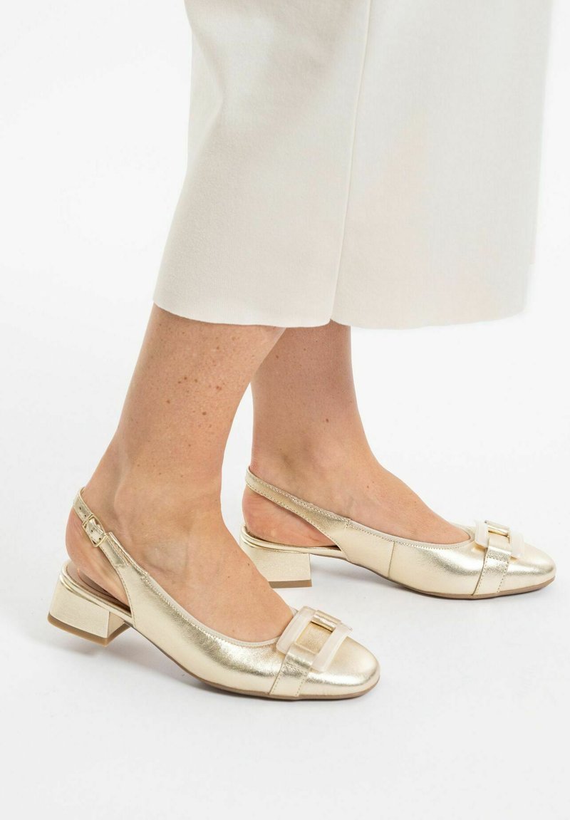 Zapatos de slingback metálicos dorados con punta cuadrada, tacón bajo y un detalle decorativo de lazo en la parte delantera. Textura de cuero suave.