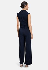 Salopette bleu marine avec un corsage ajusté, des manches courtes et un col. Présente un détail froncé au dos et un pantalon à jambes larges.