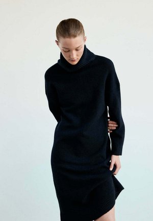 Frau mit Dutt, die ein schwarzes langärmliges Rollkragen-Strickkleid trägt, schaut nach unten und hält mit einer Hand den Saum des Kleides.