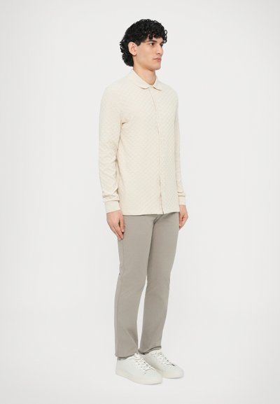 Κρεμ μακρυμάνικο πουκάμισο με υφή και γιακά, σε συνδυασμό με στενά ανοιχτό γκρι παντελόνια και λευκά αθλητικά παπούτσια, εμφανιζόμενο σε μια όρθια φιγούρα.