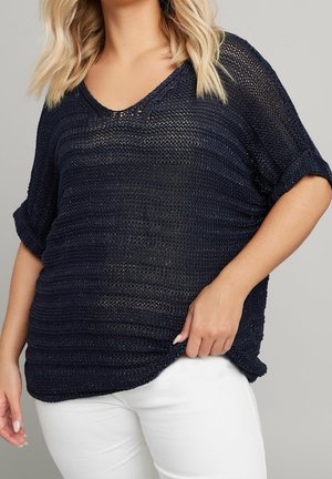 Femme portant un haut en maille noir ample à manches courtes avec des revers aux manches et un pantalon blanc sur un fond uni.