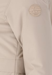 Beige takki, jossa on sileä kangas, näkyvät sauman yksityiskohdat ja pyöreä logo-merkki ylävarressa, jossa on geometrisia muotoiluelementtejä.