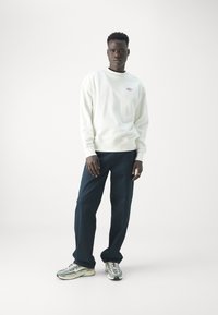 Sudadera blanca con logo, pantalones azul marino y zapatillas con estampado. El modelo se encuentra de pie contra un fondo blanco, mostrando un ajuste relajado y un estilo casual.