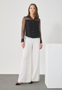 Μαύρη διάφανη μπλούZA με κουμπιά μπροστά και γιακά, συνδυασμένη με λευκό παντελόνι φαρδύ και μαύρες γόβες με μυτερή μύτη.