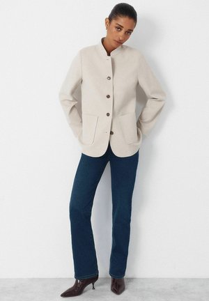 Femme portant une veste beige boutonnée avec des poches plaquées, un jean droit bleu foncé, et des bottines à talons pointues marron foncé, debout contre un mur blanc.