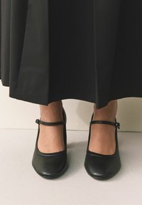 Pantofi cu toc Mary Jane din piele neagră, cu vârf rotund, baretă la gleznă și textură netedă, asortați cu o fustă neagră fluidă.