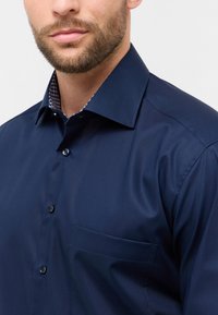 Camicia da uomo color navy con colletto a punta, chiusura frontale con bottoni, taschino discreto e dettaglio a motivo all'interno del colletto, realizzata in tessuto liscio.