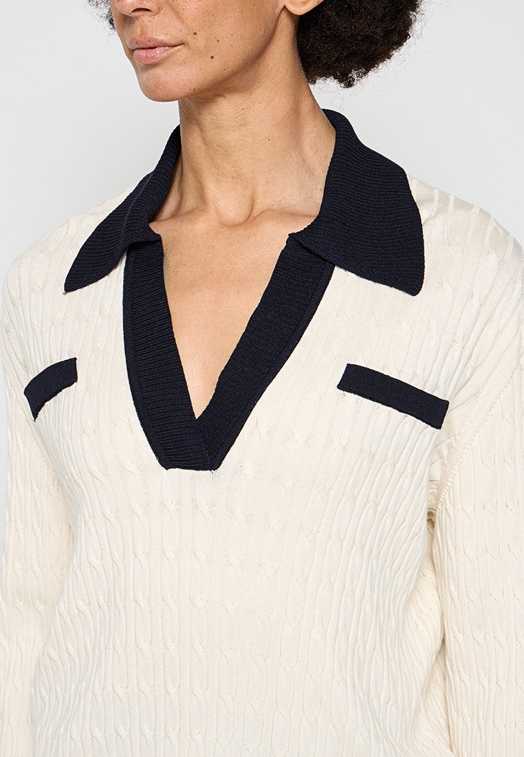 Maglione lavorato a maglia con corpo crema, colletto a coste blu navy e tasche. Presenta un motivo testurizzato e un design a scollo a V profondo.