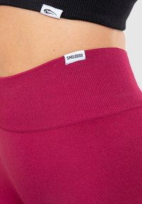 Nämä syvän vaaleanpunaiset, korkea vyötäröiset leggingsit ovat tiukasti istuvat ja niissä on ribbireunus. Tägeissä lukee "SMILODOX" ja osittain näkyvissä on musta toppi.