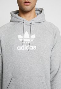 Světle šedá mikina s kapucí vyrobená z měkkého materiálu, s centrálním bílým vyšívaným logem adidas a klokankou. Šňůrka na kapuci.