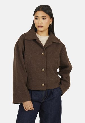 Vrouw draagt een bruine wollen jas met knopen en brede kraag, en een donkerblauwe spijkerbroek. Ze staat met één hand in haar zak en kijkt opzij.