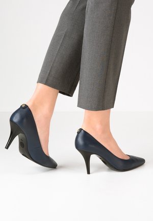 Pieds portant des escarpins en cuir bleu marine associés à un pantalon gris retroussé, debout sur un sol blanc devant un fond uni.