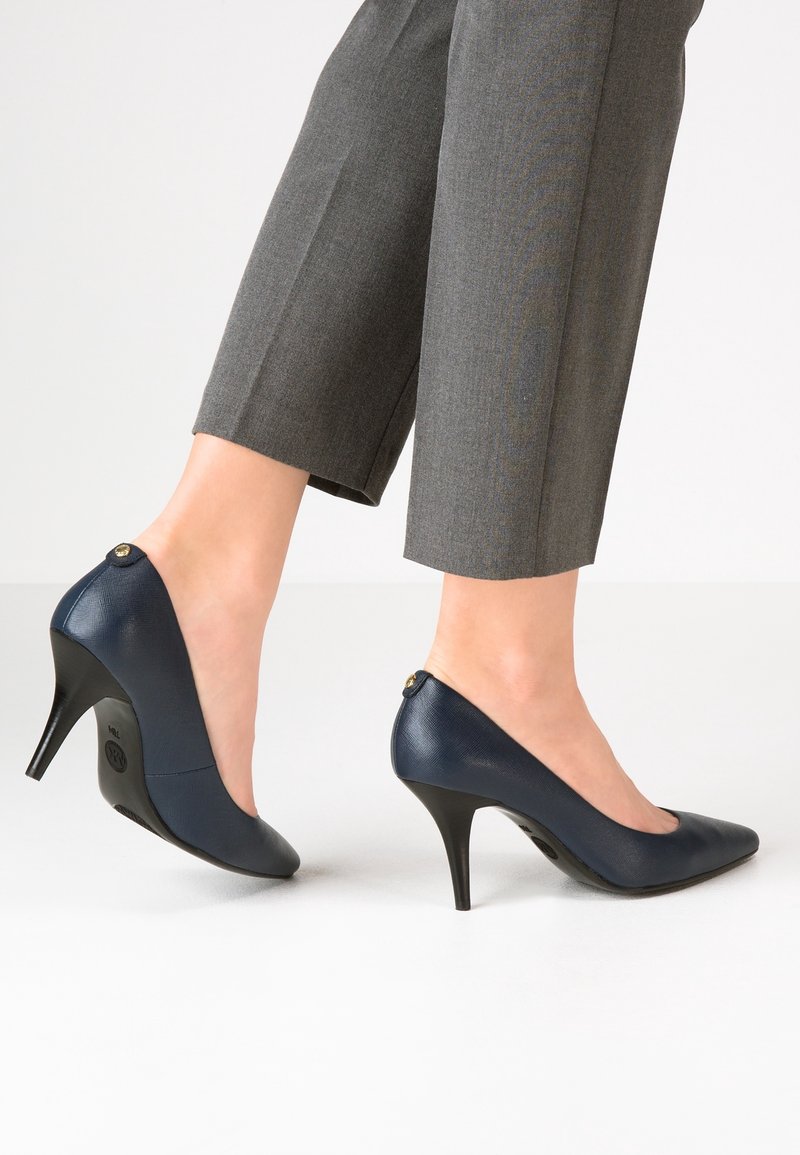 Pieds portant des escarpins en cuir bleu marine associés à un pantalon gris retroussé, debout sur un sol blanc devant un fond uni.
