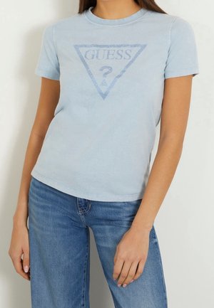 Kobieta ubrana w jasnoniebieską koszulkę z krótkim rękawem z logo "GUESS USA" i niebieskie jeansy, stojąca na tle jednolitego, jasnego tła.