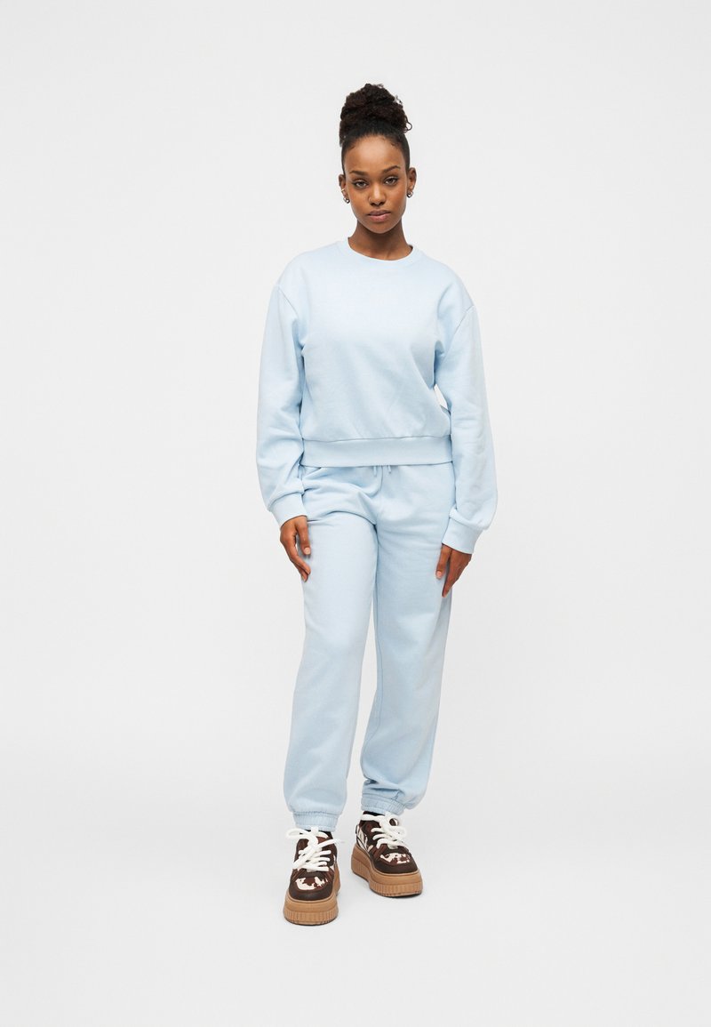 Γυναίκα που στέκεται με ανοιχτογάλαζο φούτερ και φόρμες, φορώντας καφέ και λευκά παπούτσια πλατφόρμα, μπροστά σε απλό λευκό φόντο.