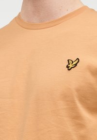 Ανοιχτό πορτοκαλί βαμβακερό t-shirt με στρογγυλή λαιμόκοψη και μικρό κεντημένο λογότυπο πουλιού σε μαύρο και κίτρινο χρώμα στο αριστερό στήθος. Ομαλή υφή.