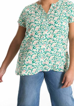 Person iført en kortærmet grøn blomsterbluse med pink og hvide blomster parret med blå jeans, stående med afslappede arme.