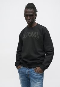 Μαύρο φούτερ με το λογότυπο "DIESEL" σε κεντήματα με αντίθεση, στρογγυλή λαιμόκοψη, ριμπ τελειώματα στα μανίκια, συνδυασμένο με ανοιχτόχρωμο τζιν παντελόνι.