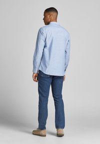 Camisa de botón azul claro de manga larga, con un dobladillo curvado, combinada con unos vaqueros azul oscuro y zapatos beiges. Vista trasera mostrada.