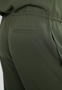 Pantalon vert olive avec taille élastique et poche arrière fendue horizontalement du côté droit, présenté en vue rapprochée.